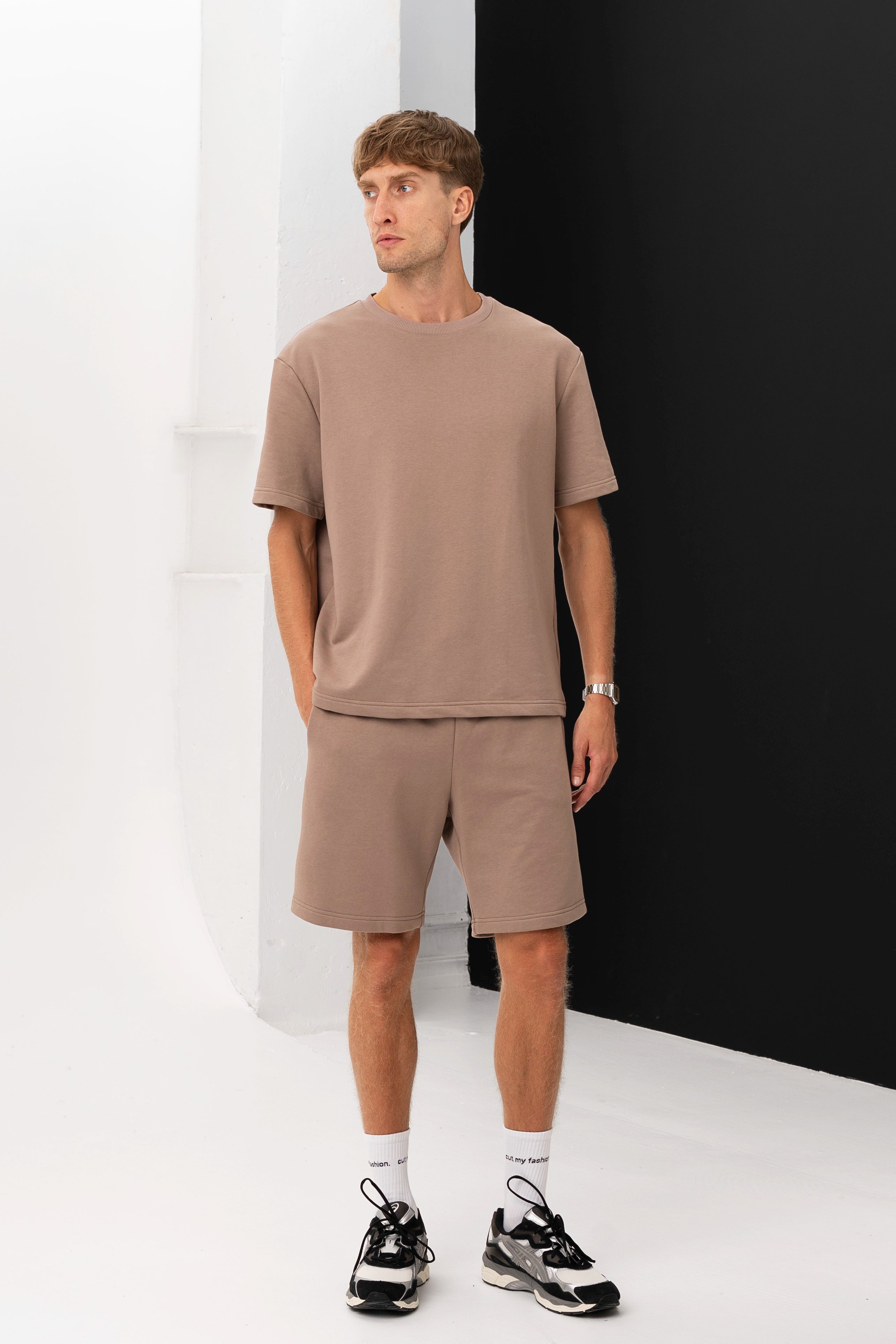 A model in a brown T-shirt and shorts poses in a minimalist studio with his hands in his pockets