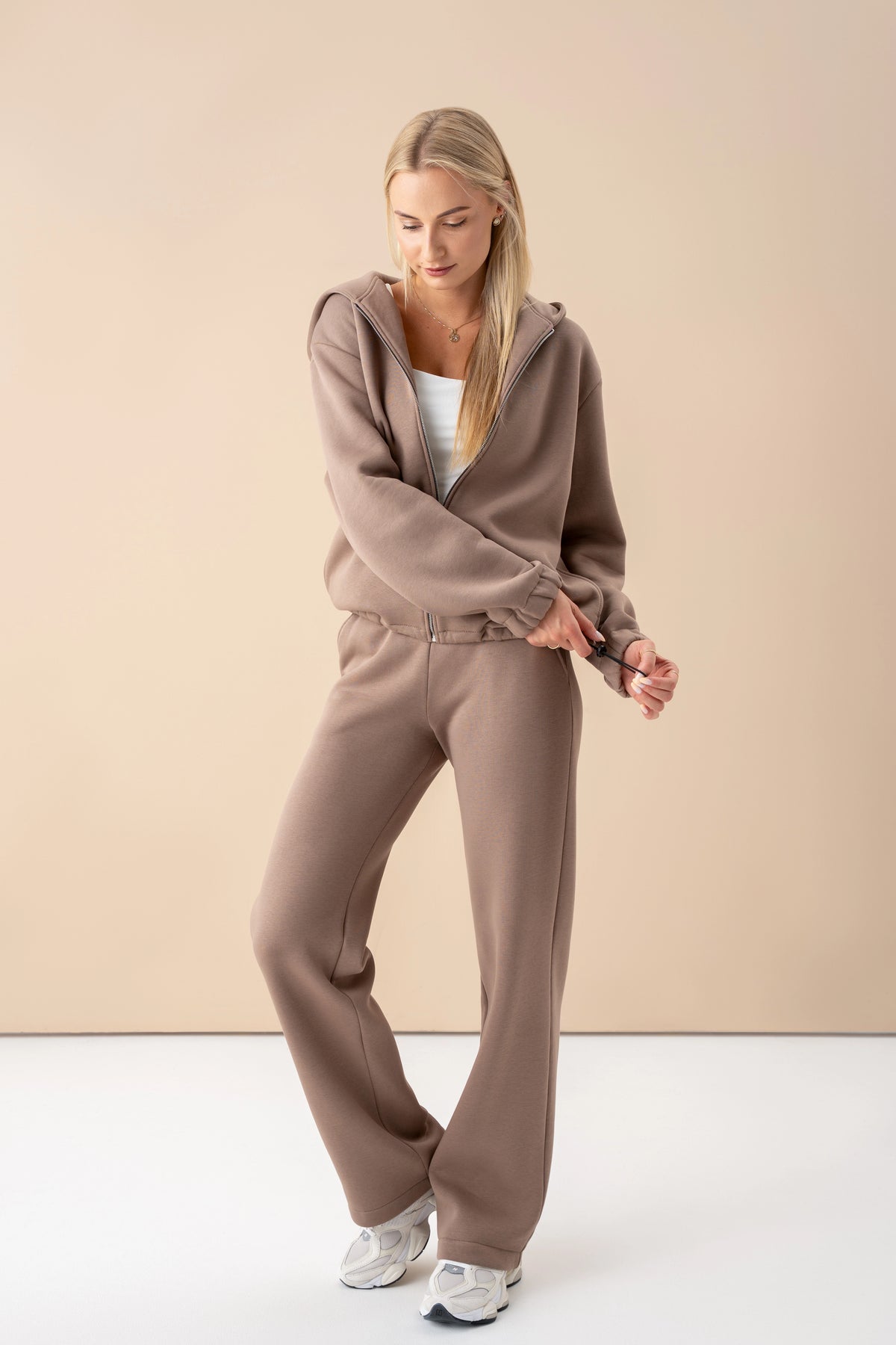 model wearing a latte-colored tracksuit with a zip-up sweatshirt, standing in the studio, looking down while adjusting the width of the sweatshirt