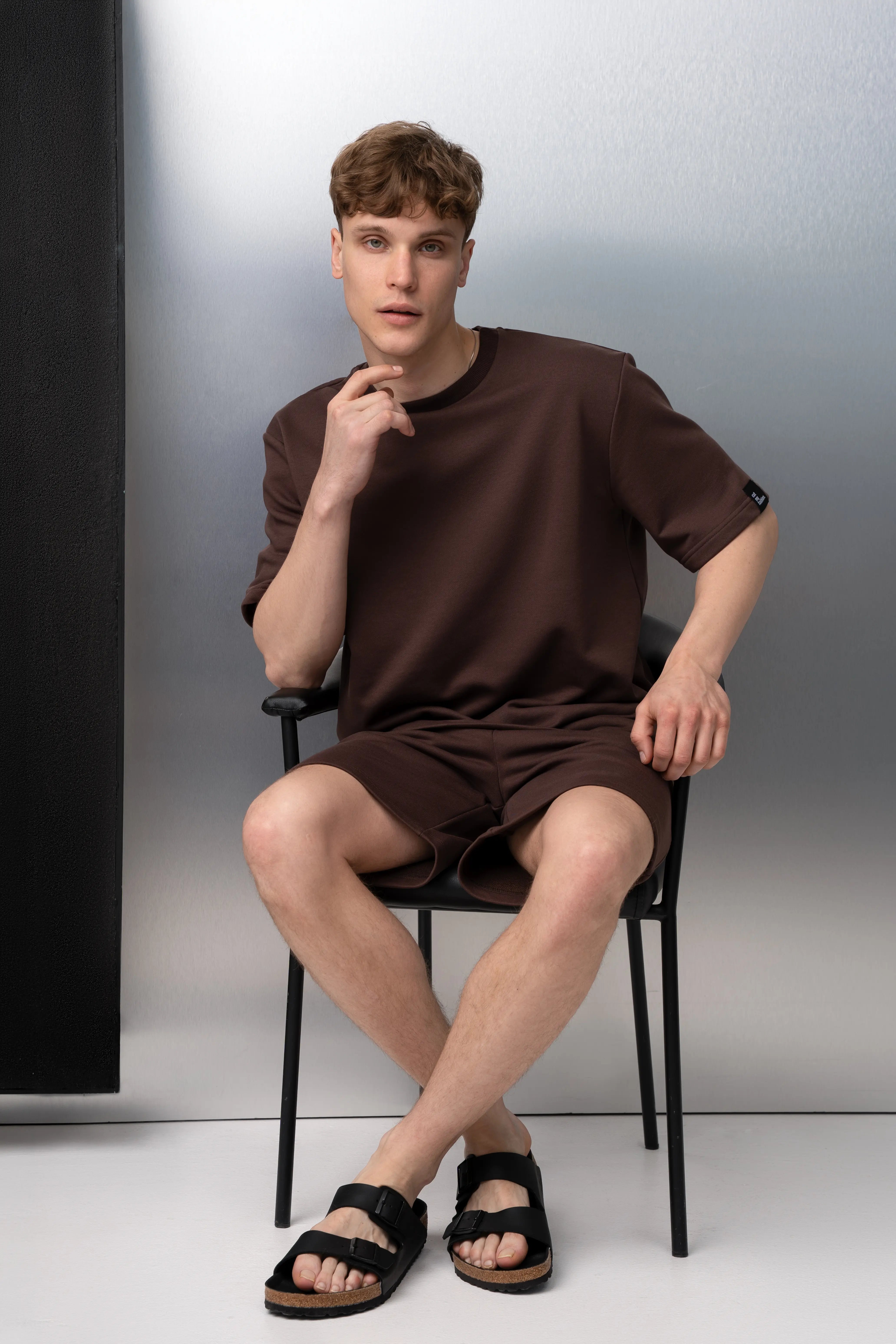 Vyras sėdi vilkėdamas rudos spalvos kostiumėlį iš be pūkelio audinio su oversized marškinėliais ir šortais