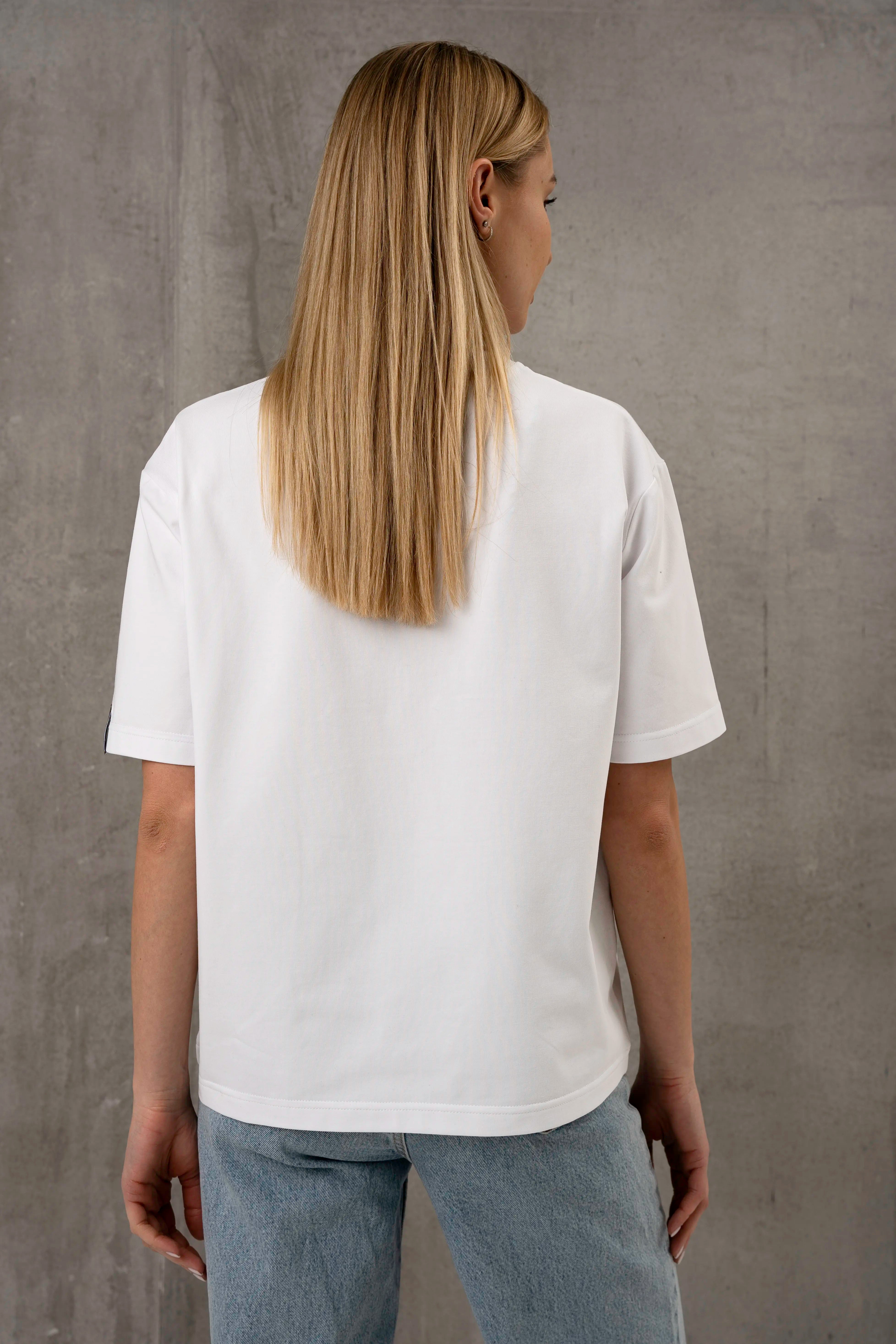 A woman is wearing a white, oversized short-sleeved T-shirt and light-colored jeans; she is shown from behind against a gray background.