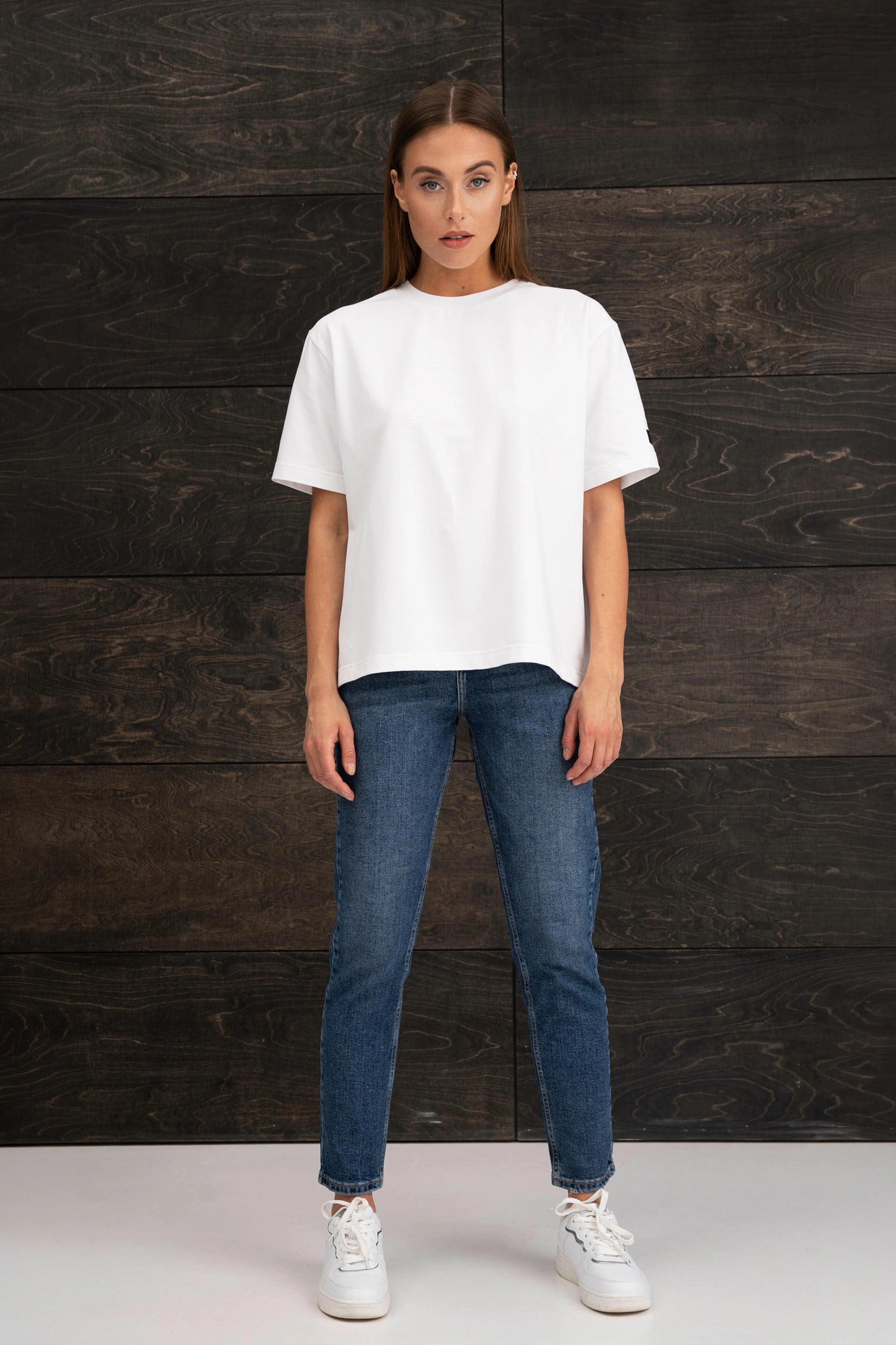 The woman is wearing a white, oversized short-sleeved T-shirt, blue jeans, and white sneakers, and is standing in front of a wall with a dark wood-grain pattern.