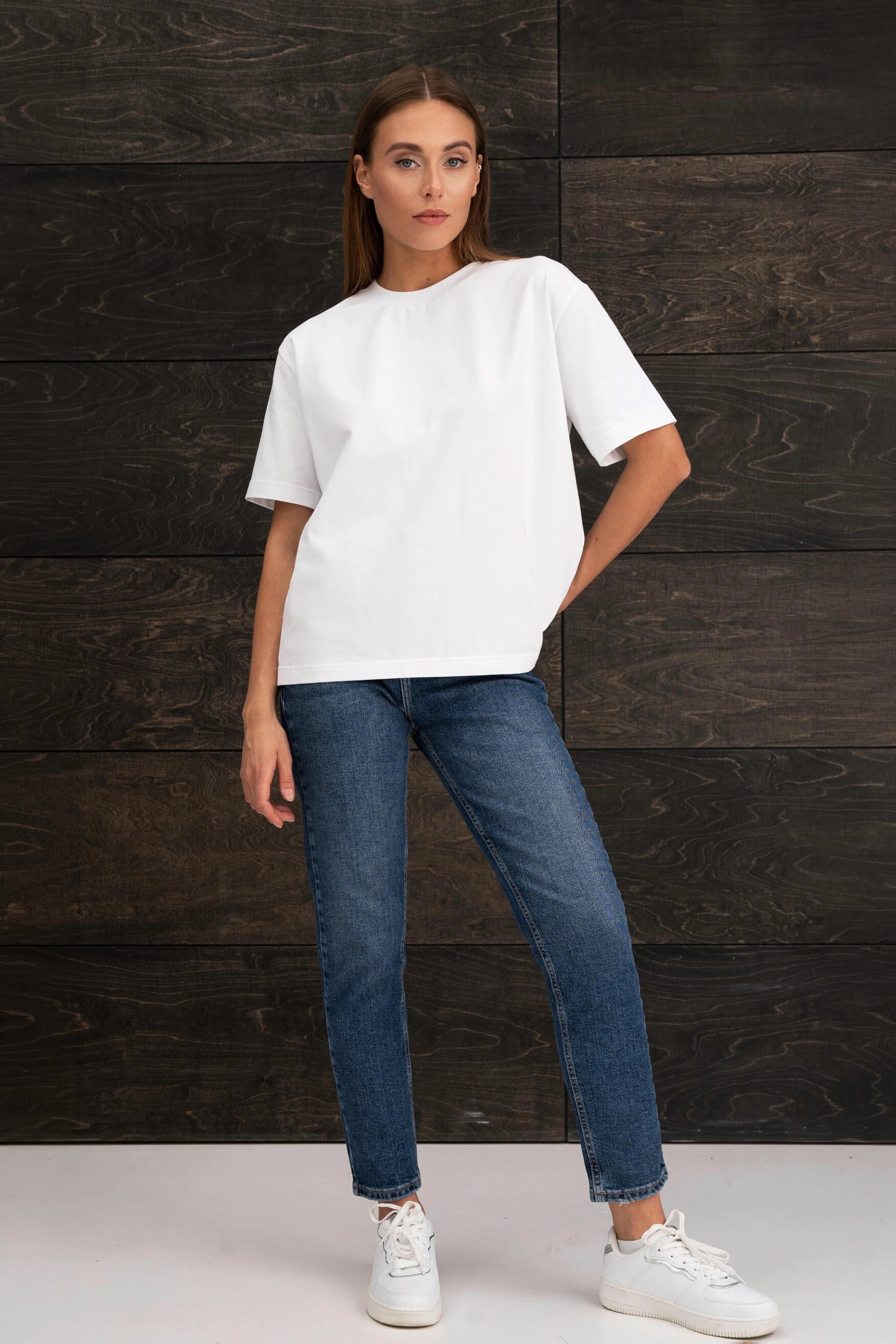 A woman is wearing a white, oversized short-sleeved T-shirt, blue jeans, and white sneakers; she is standing against a wall with a dark wood-grain pattern, her hand in her pocket.