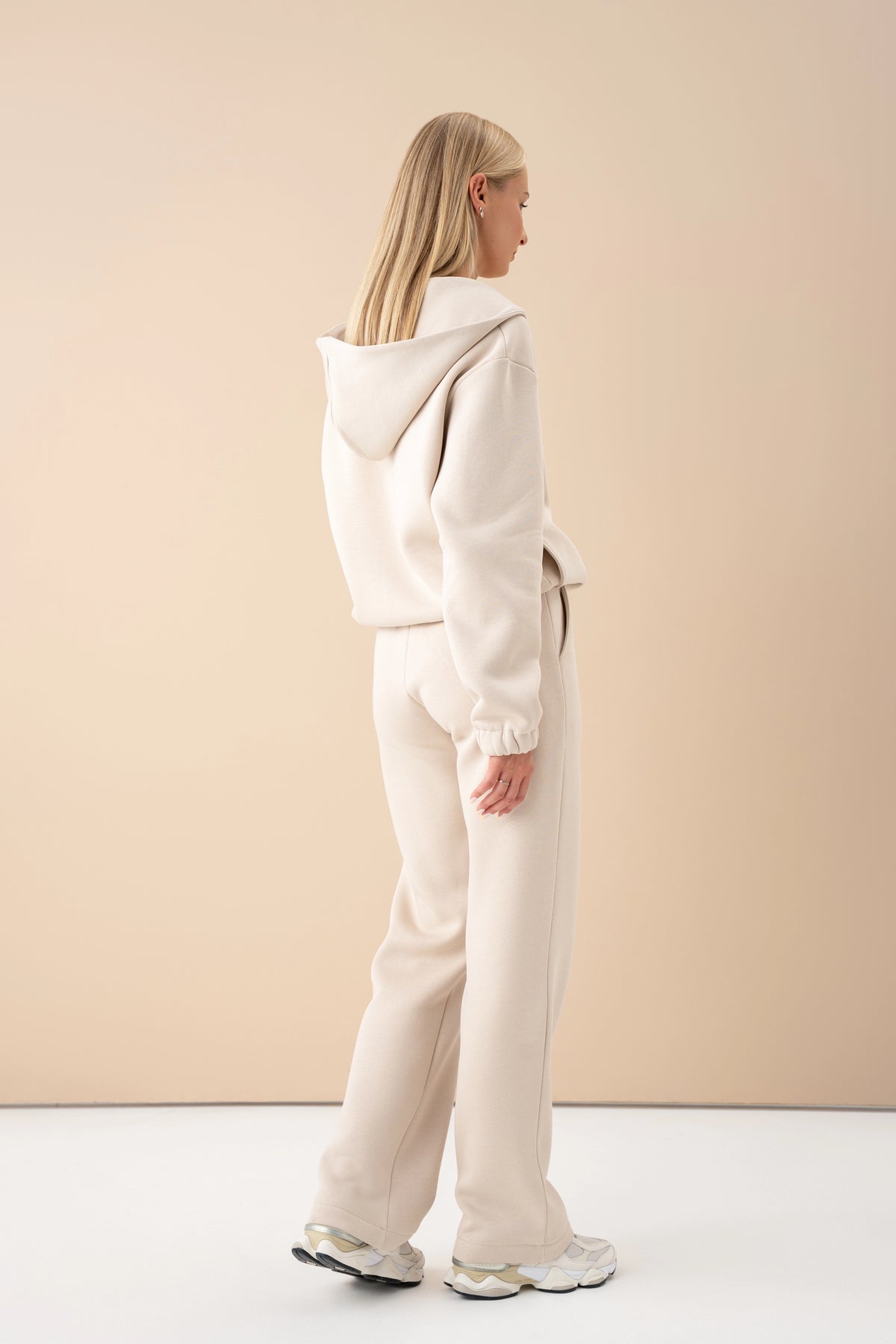 The model in a light-coloured tracksuit is turned with her back to the camera, showing the hood, with a light wall in the background