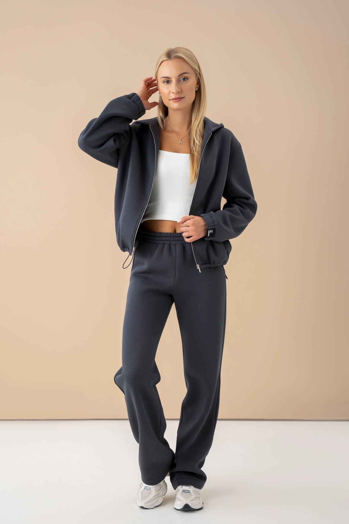 the woman is wearing an iron-colour leisure set, Made in Lithuania, posing against a light background, with one hand touching her hair and the other touching the sweatshirt