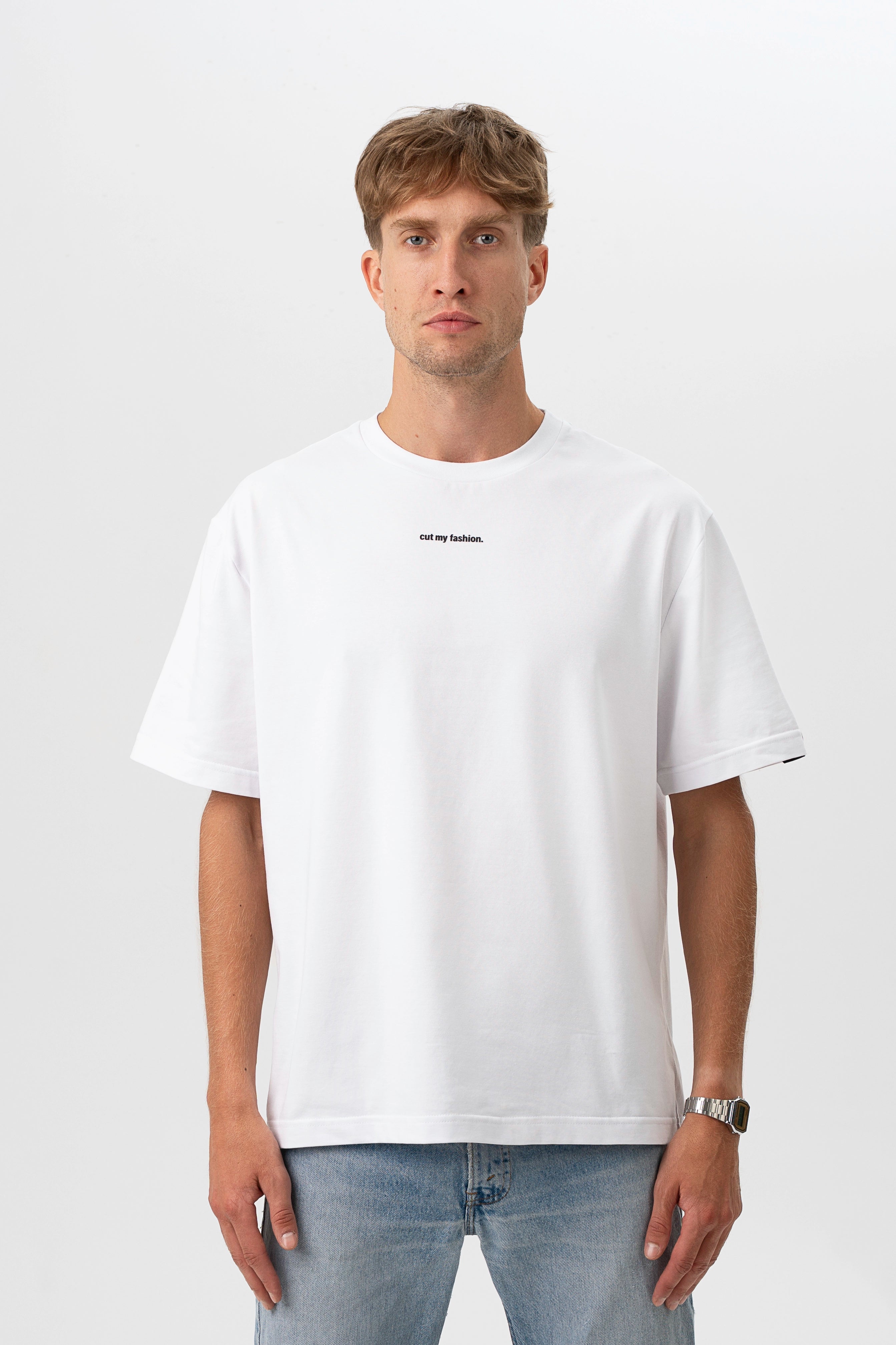 Vyras pozuoja su baltais marškinėliais „cut my fashion“ logotipu centre, neutralioje baltai pilkoje aplinkoje