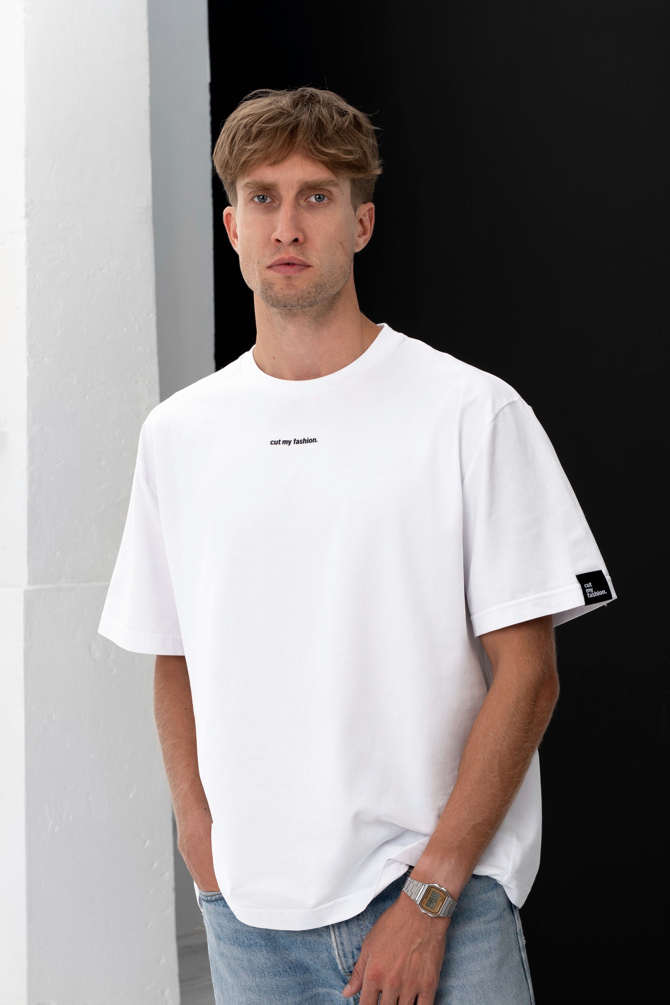 Jaunas vyras su baltais oversized marškinėliais iš organinės medvilnės ir džinsais, stovintis prieš juodą foną