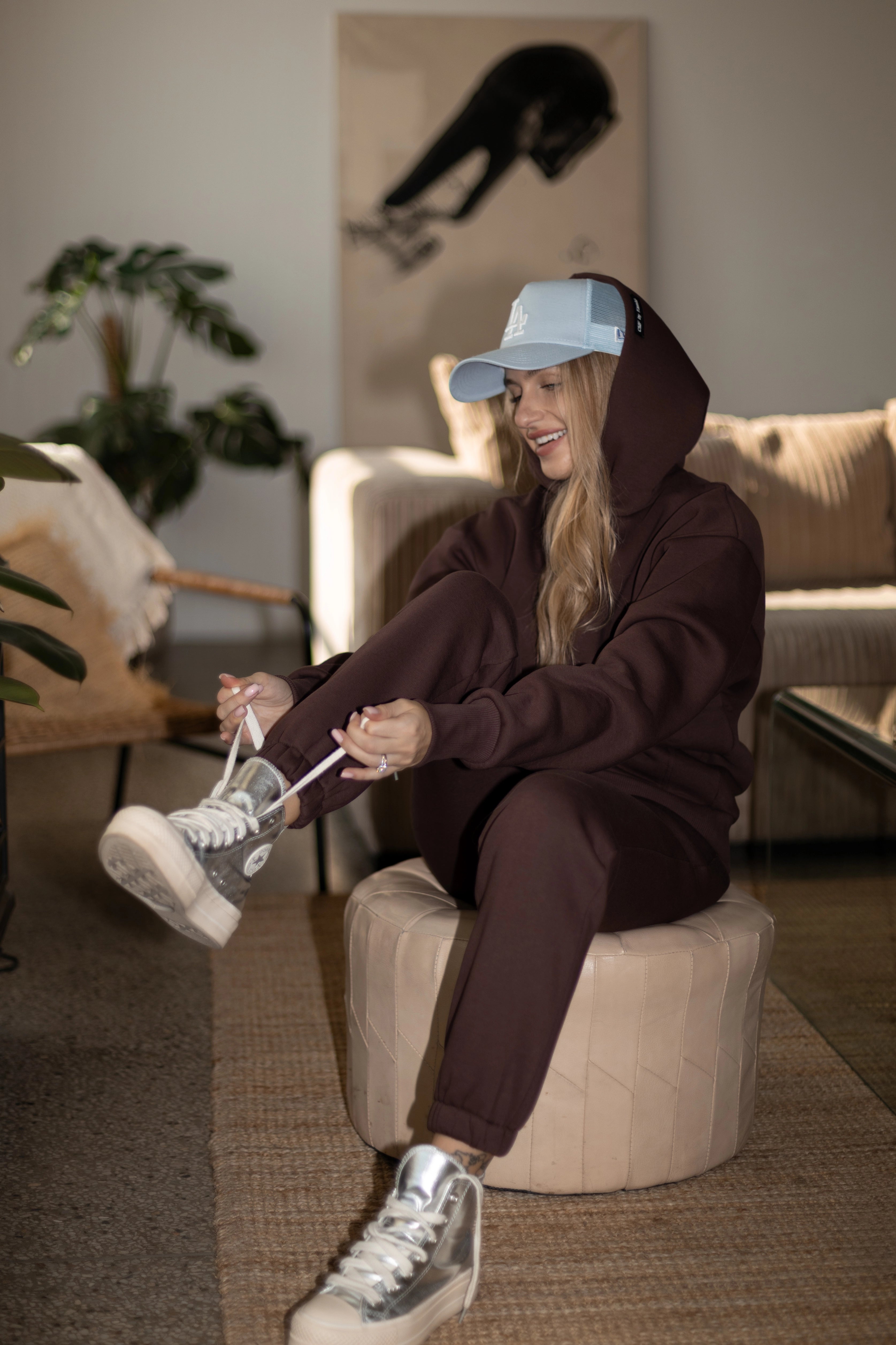 Moteris su tamsiai rudu laisvalaikio treningu sėdi ant pufiko, riša sportbačių raištelius, dėvi šviesiai mėlyną kepurę