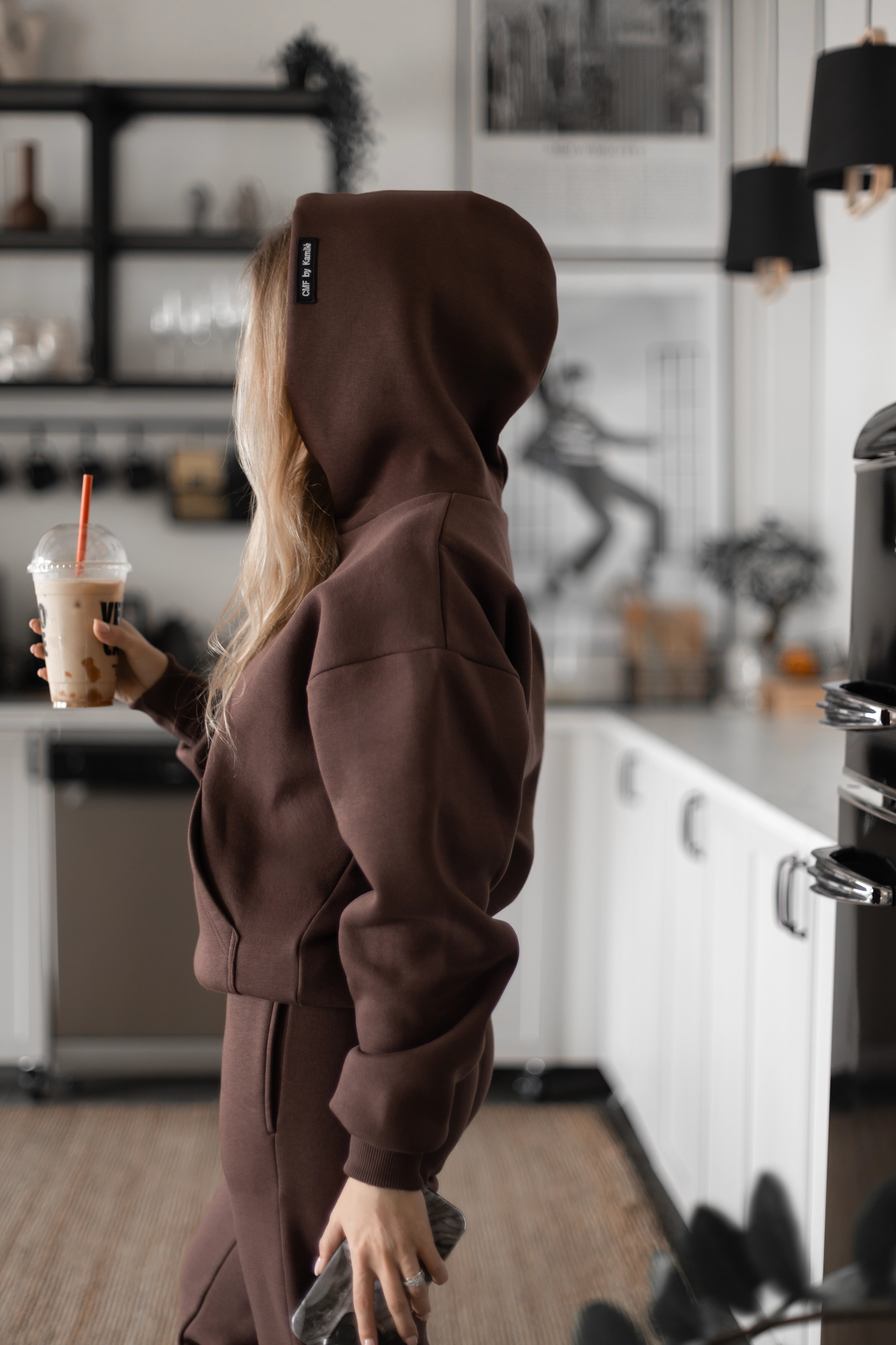 Moteris su tamsiai rudu treningu stovi modernioje virtuvėje, rankoje laiko gėrimą su šiaudeliu