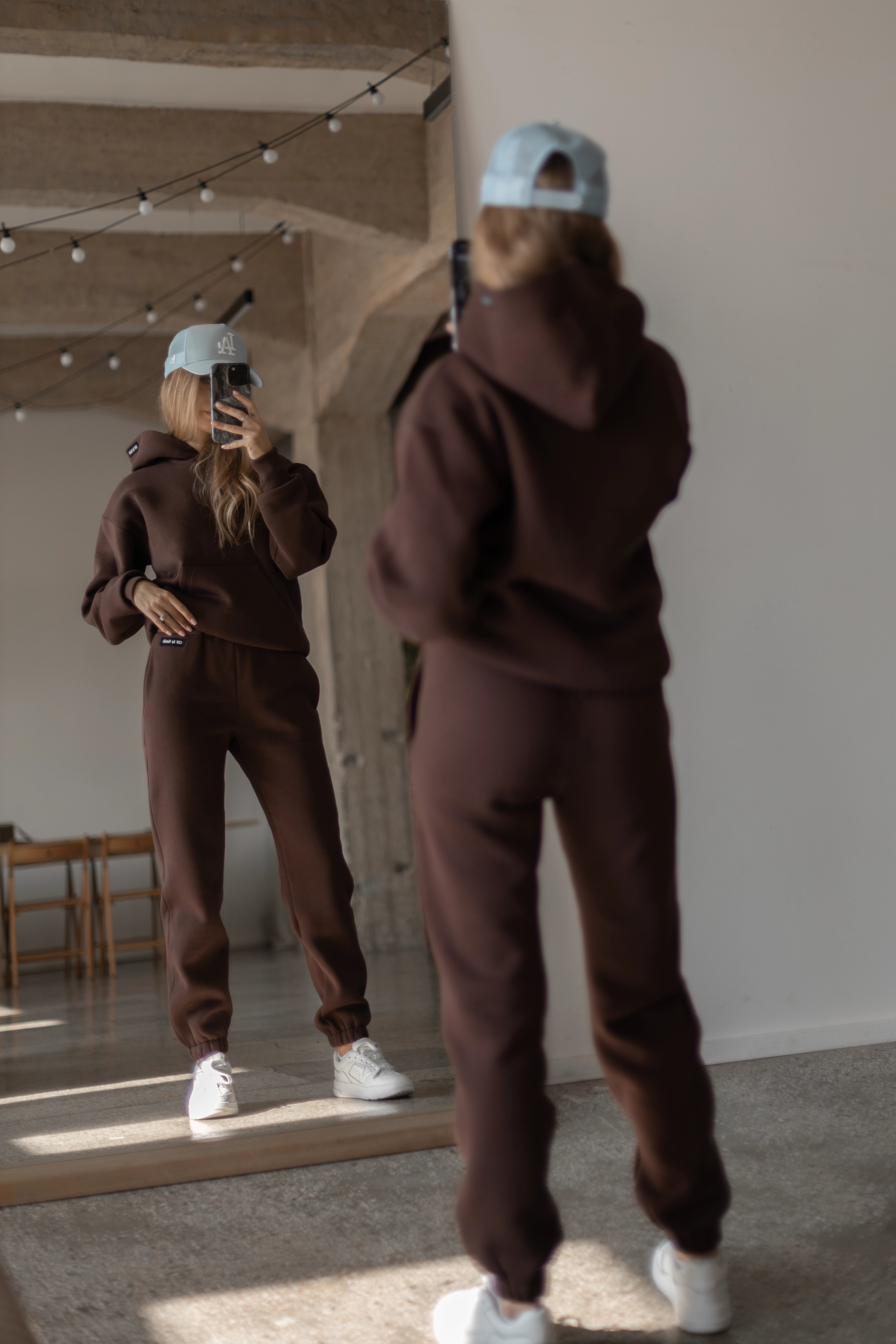 Moteris su tamsiai rudu treningu žiūri į save veidrodyje, dėvi šviesiai mėlyną kepurę ir baltus sportbačius