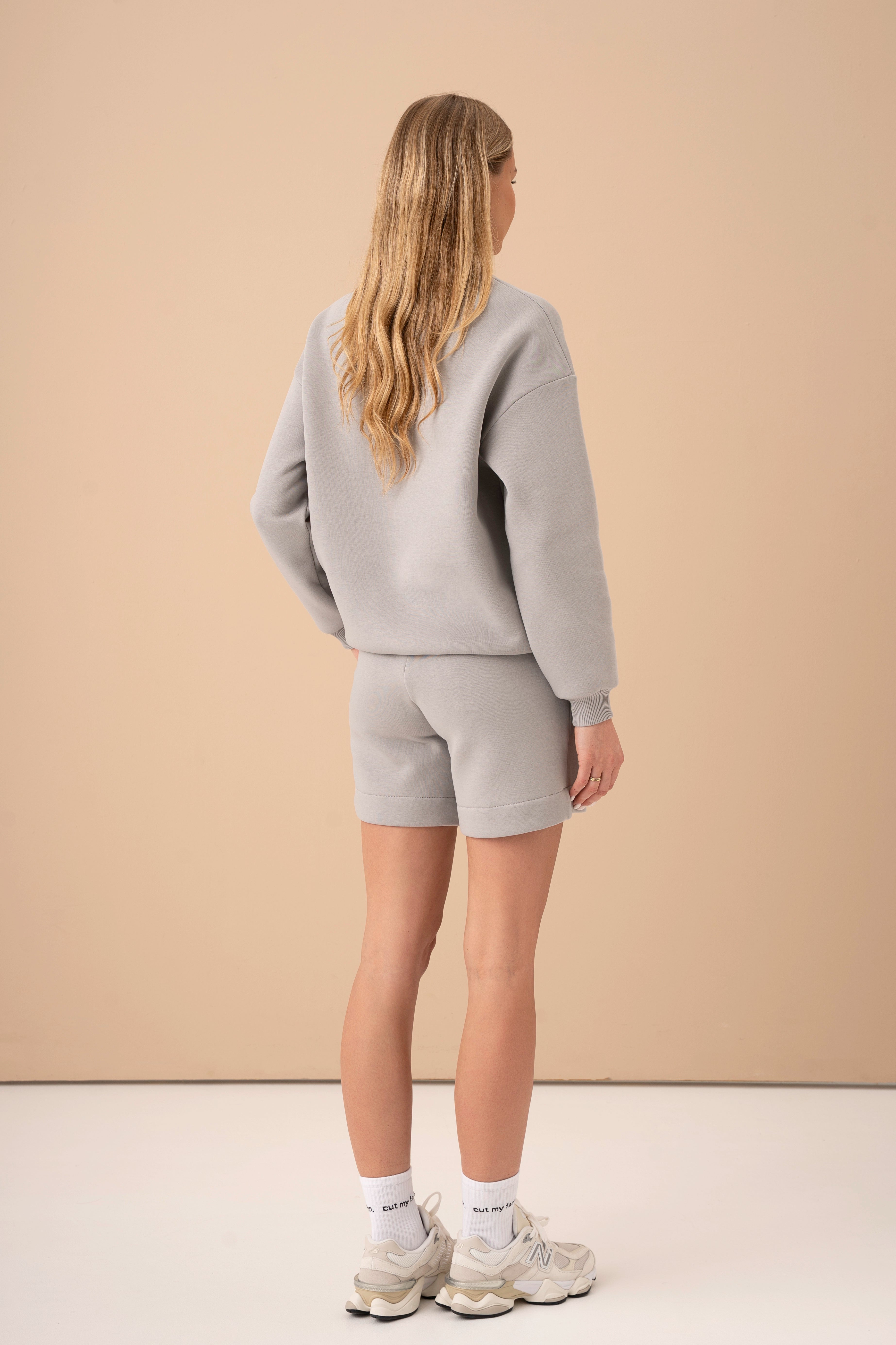 moteris su šviesiai pilku cutmyfashion laisvalaikio treningu atsisukusi nugara, matomi aukšto liemens šortai ir ilgos rankovės džemperis be kapišono, laisvalaikio treningas studijoje, pagamintas Lietuvoje