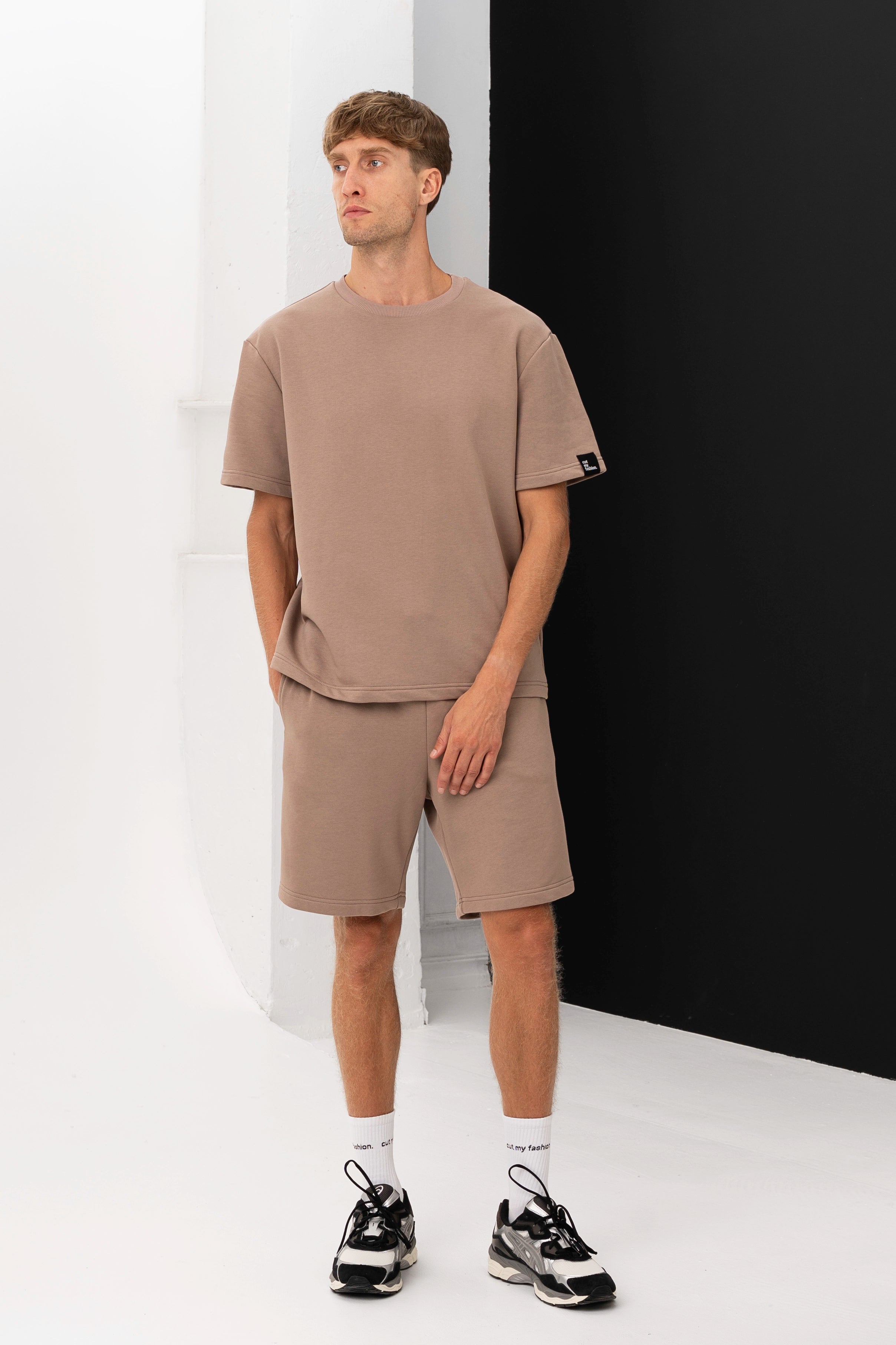 A man wears a brown T-shirt and shorts, standing by a white wall with a black detail