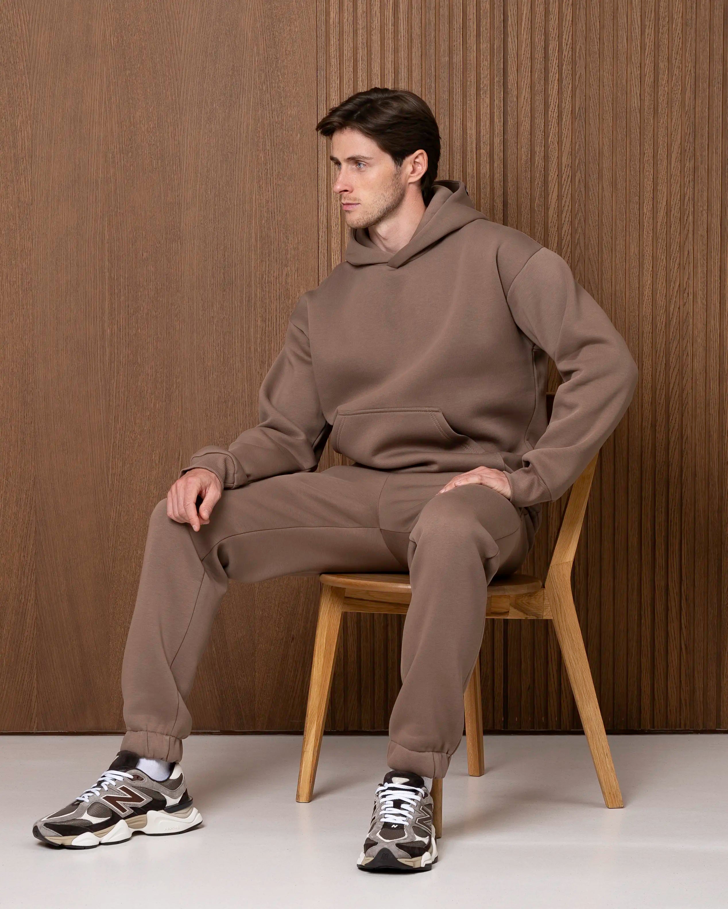 Vyras sėdi vilkėdamas smėlio spalvos treningų komplektą su gobtuvu, matomas džemperio ir kelnių kritimas.