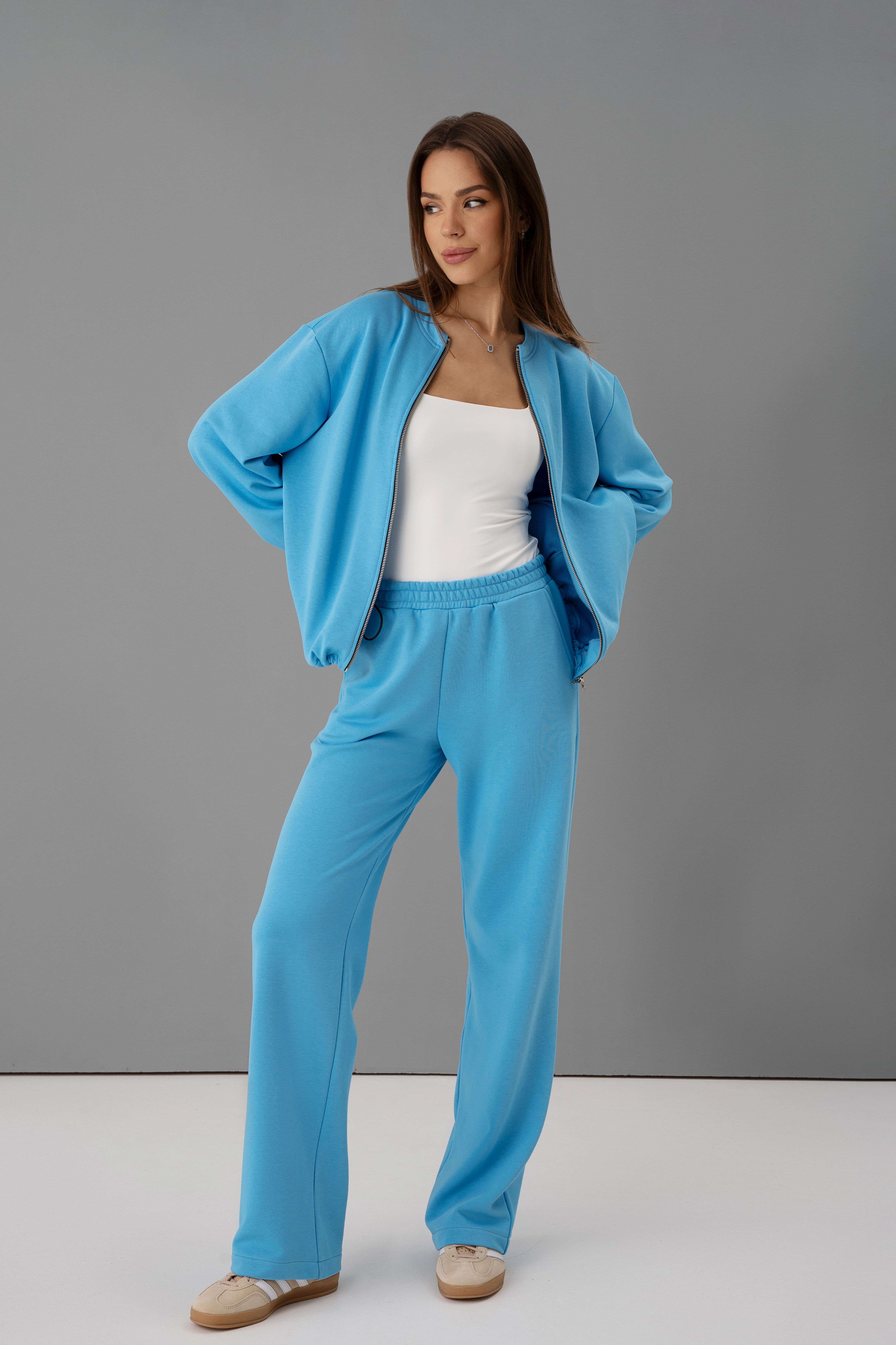 moteris su žydru užtraukiamu džemperiu ir treninginėmis kelnėmis, cutmyfashion pagaminta Lietuvoje, pozuoja šviesiame fone