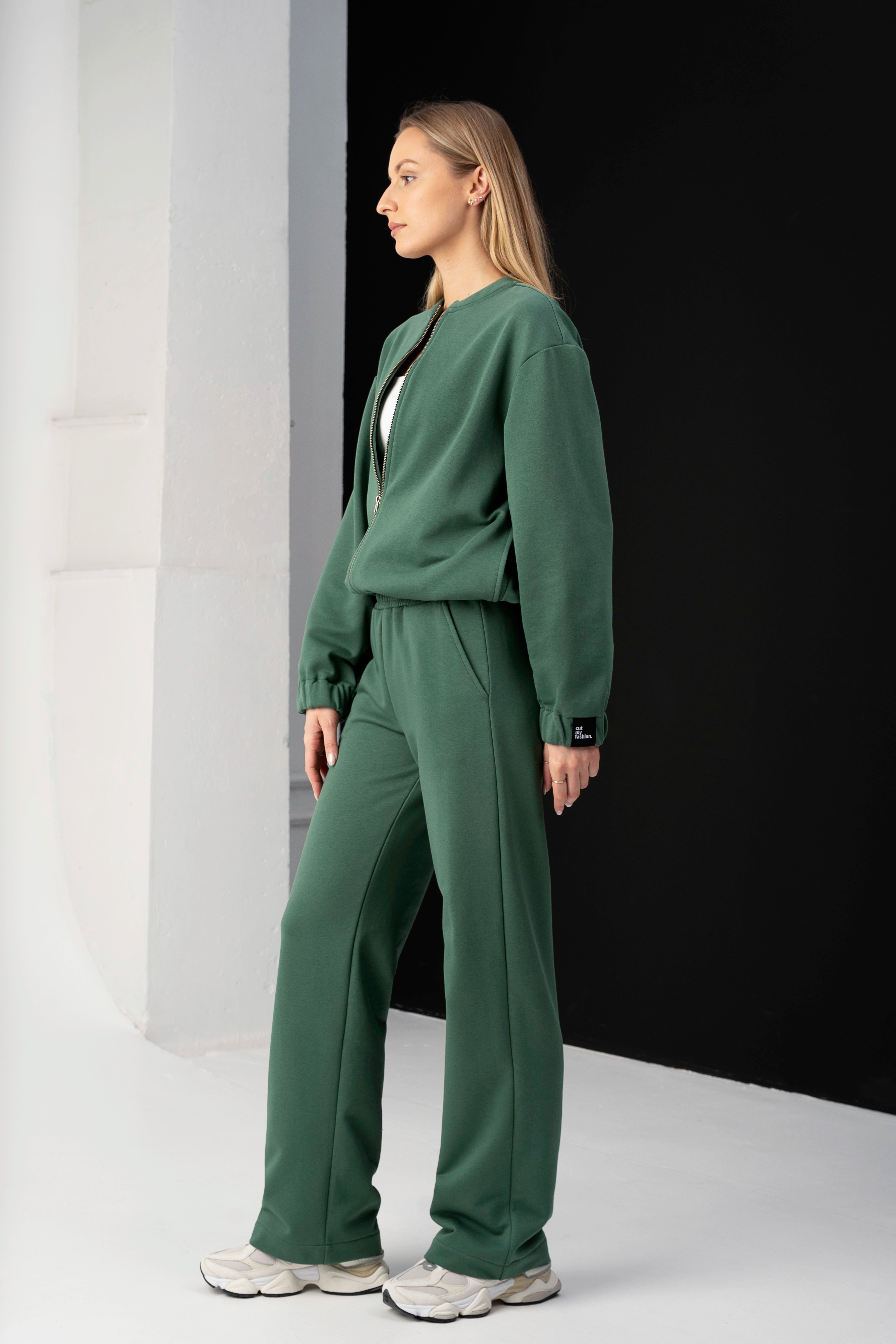 moteris dėvi žalius cutmyfashion treningus mėja, stovi profiliu studijos fone