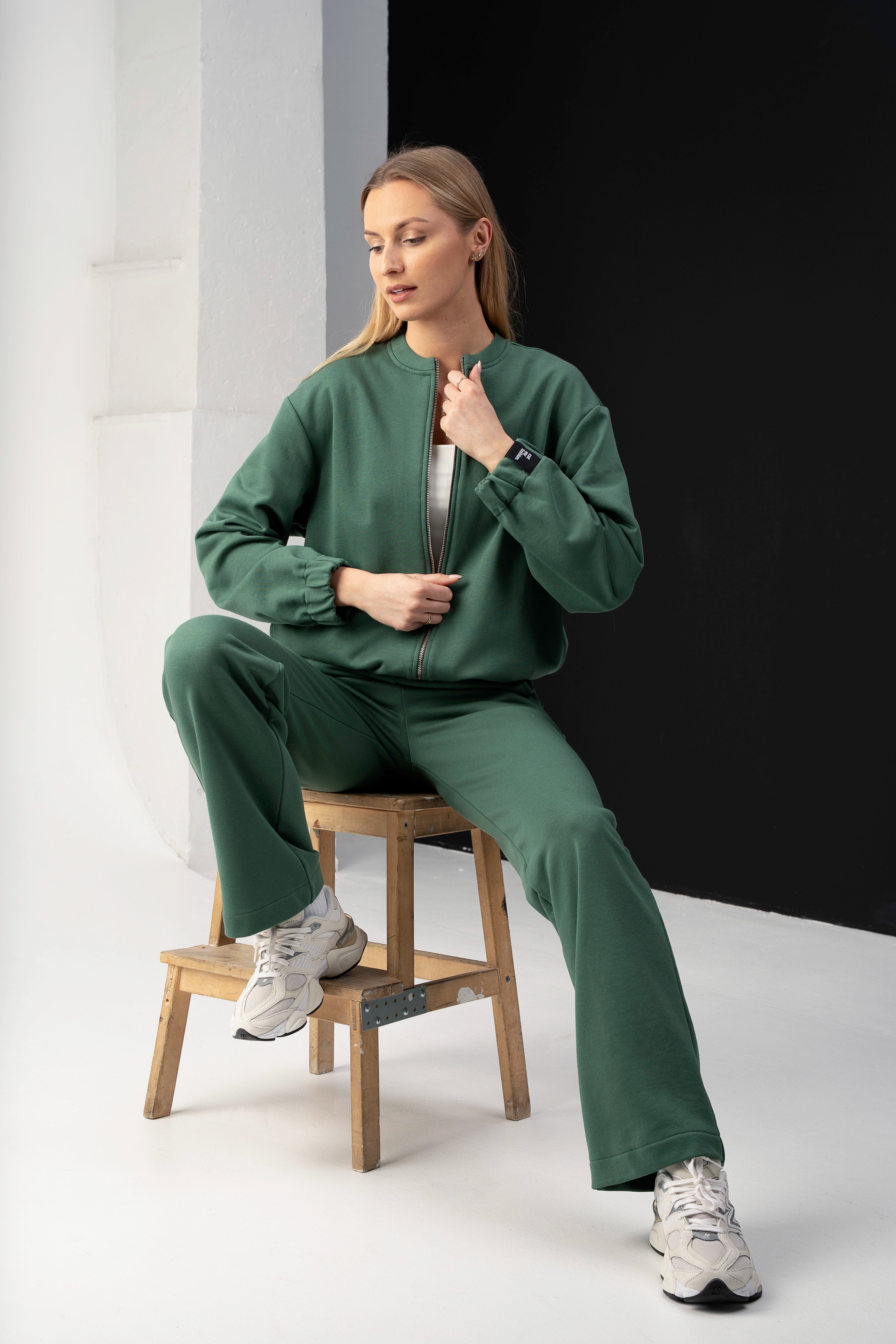 modelis su žaliais cutmyfashion treningais mėja, atsisėdusi ant medinės kėdutės studijoje, atsega džemperį