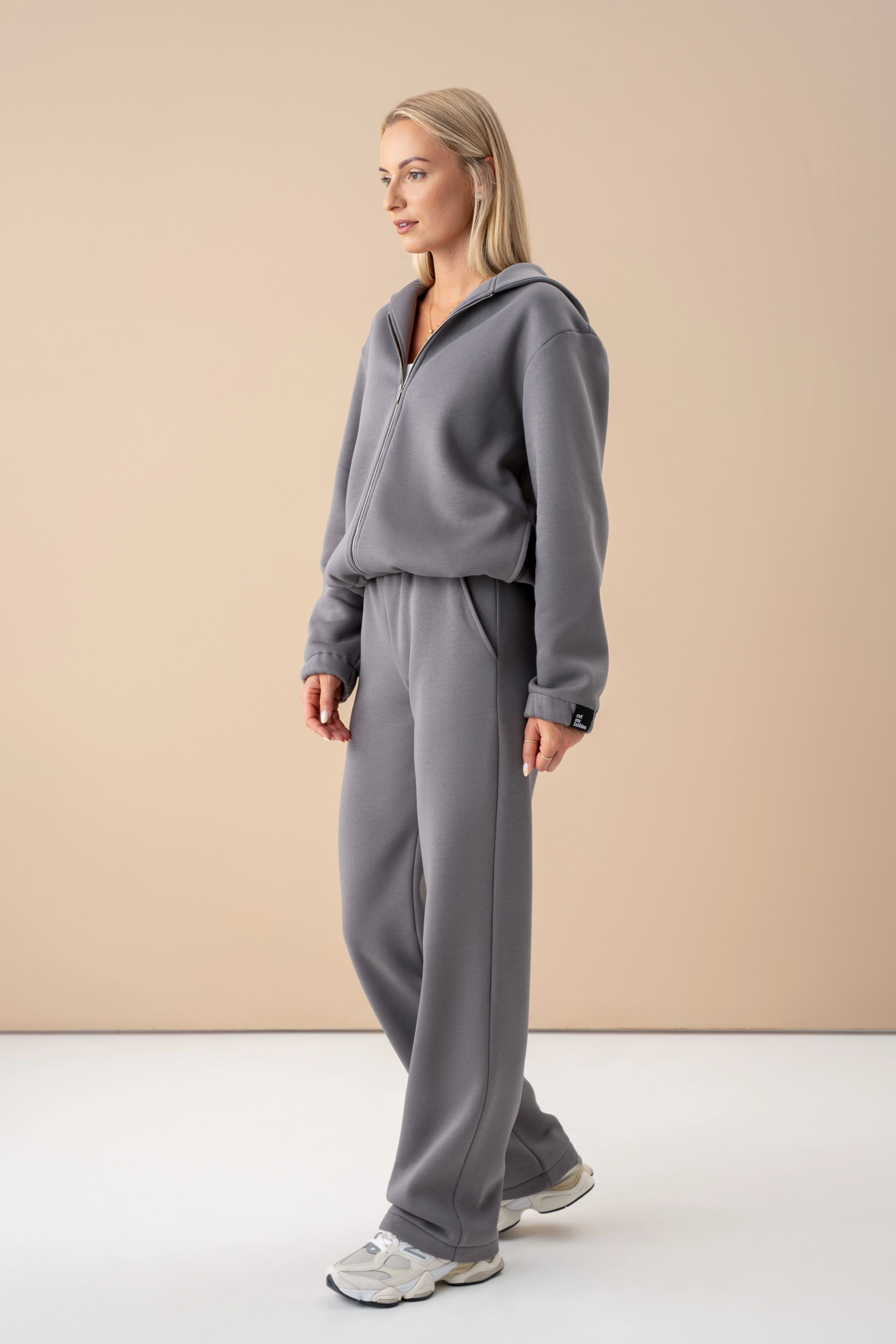 moteris su pilkos spalvos cutmyfashion treningais pasisukusi šonu, pozuoja šviesiame studijos fone