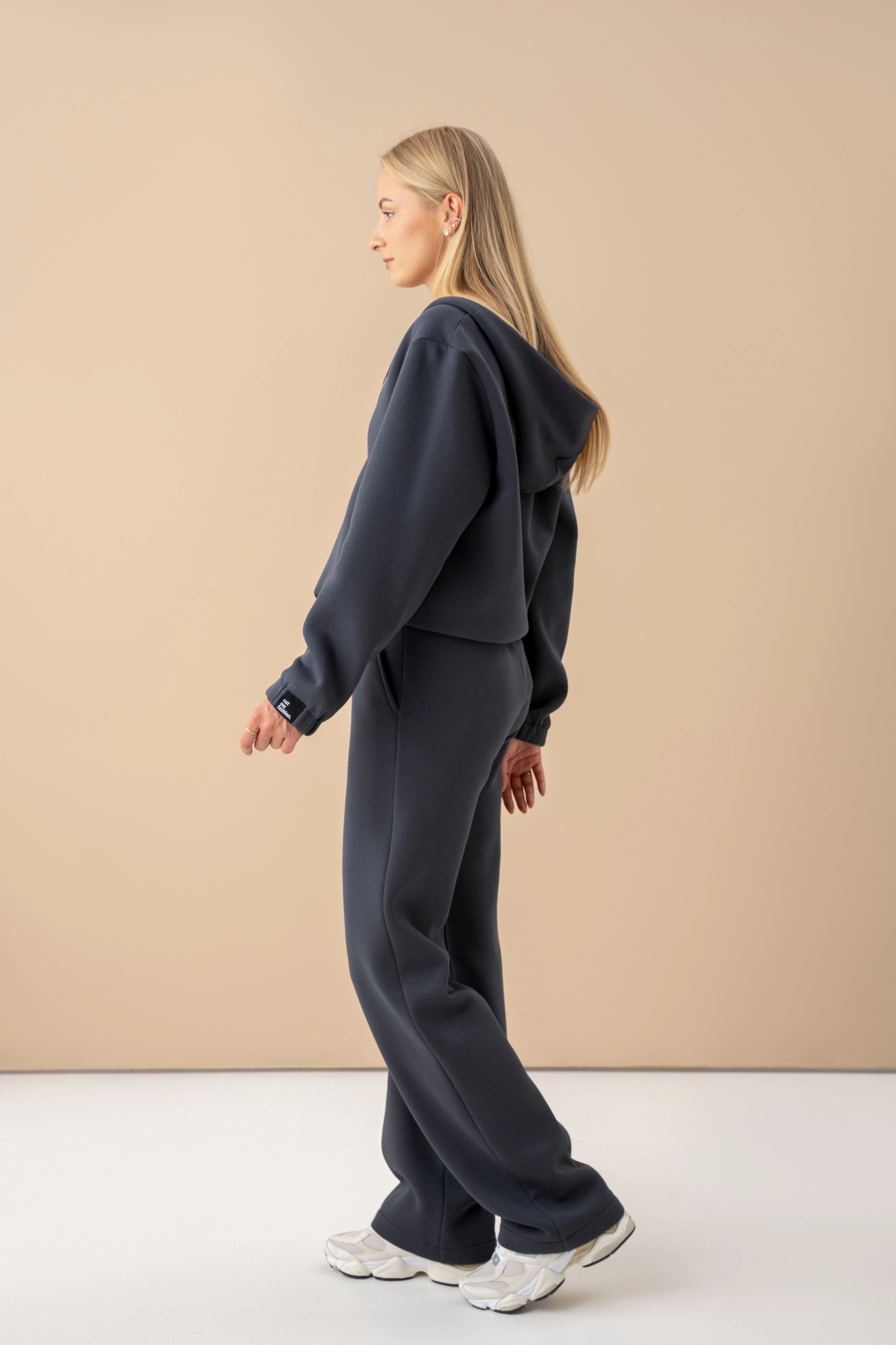 modelis su tamsiai pilku cutmyfashion laisvalaikio treningais, stovi šviesiame studijos fone pasisukusi šonu