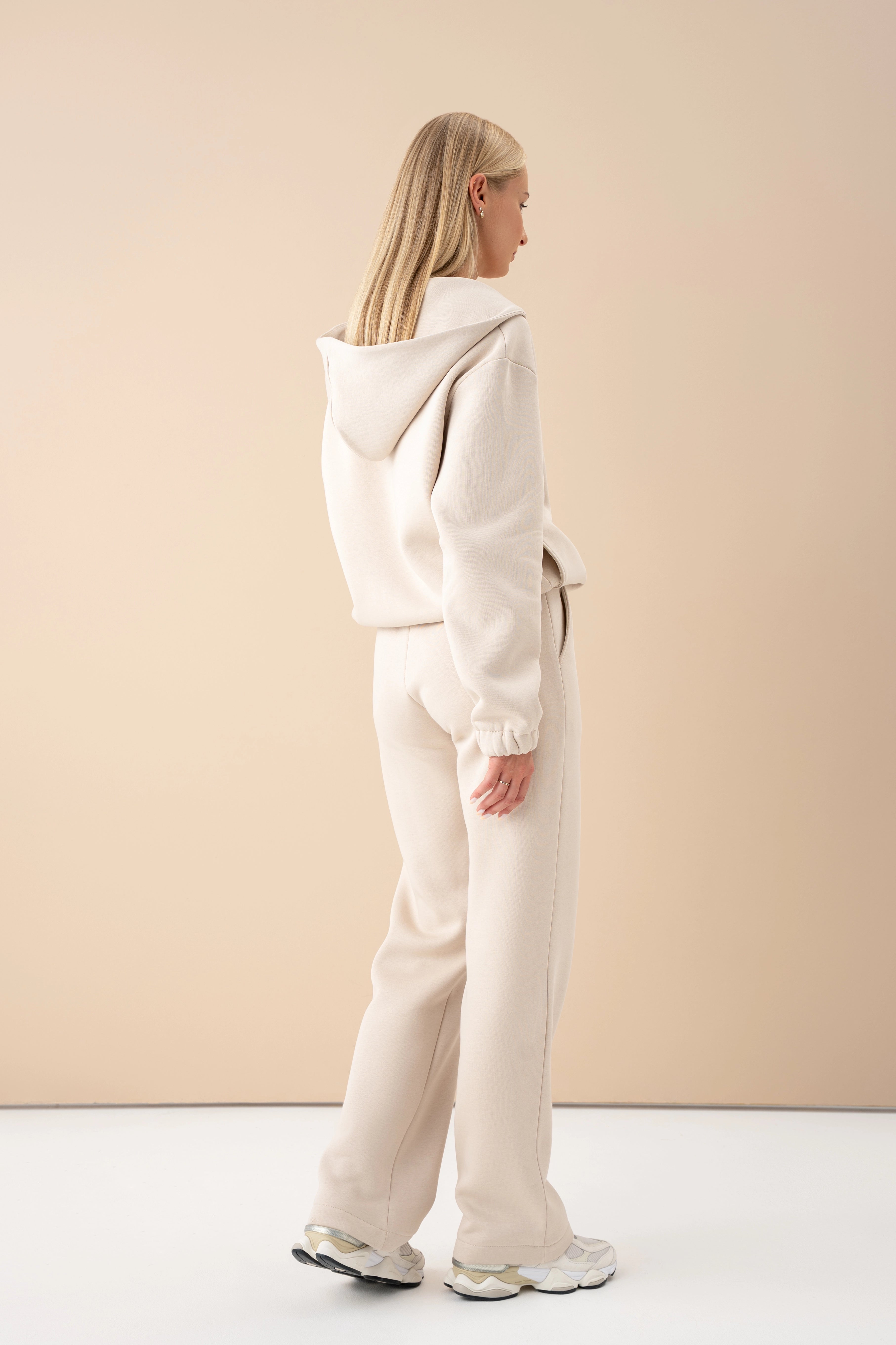 The model in a light-coloured tracksuit is turned with her back to the camera, showing the hood, with a light wall in the background