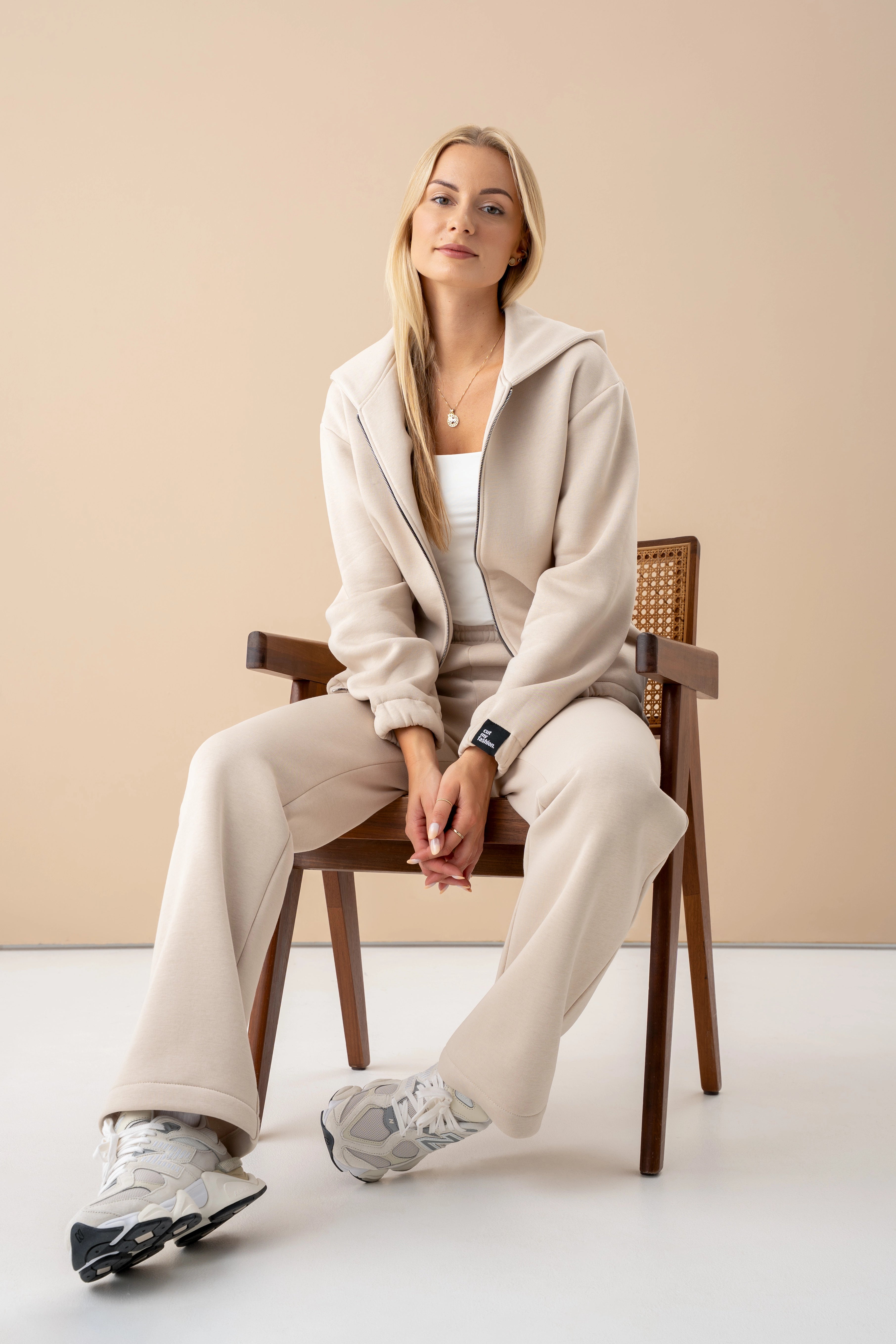 A woman wearing cutmyfashion tracksuits is sitting on a chair, wearing a zip-up sweatshirt, looking into the camera