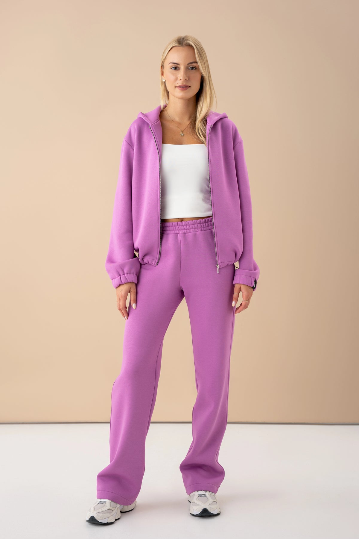 The model is wearing a pastel pink tracksuit set with a zip-up sweater, looking into the lens against a light background