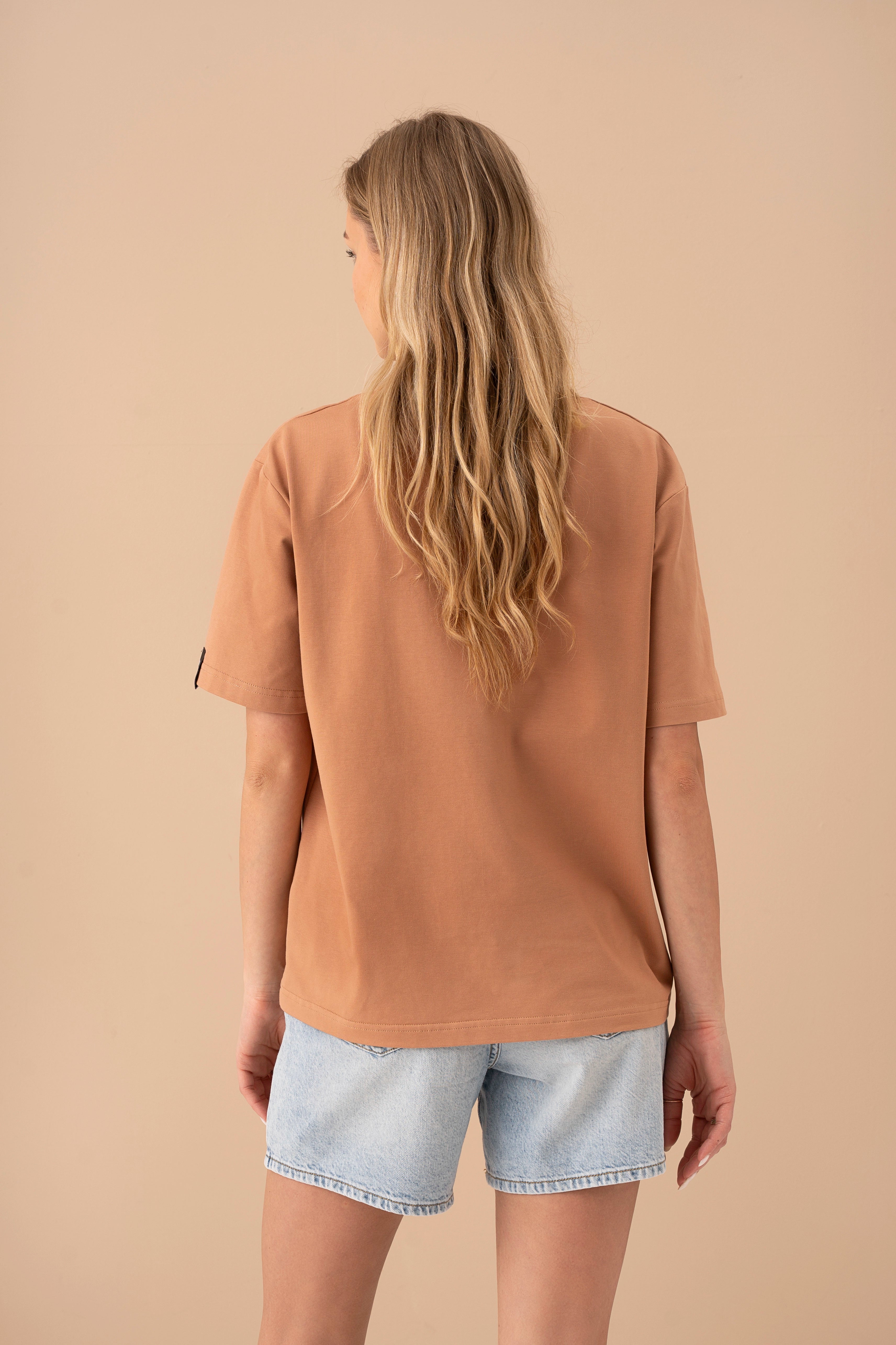 the model wearing light denim shorts and an oversized mocha T-shirt is turned with her back to the camera, studio background