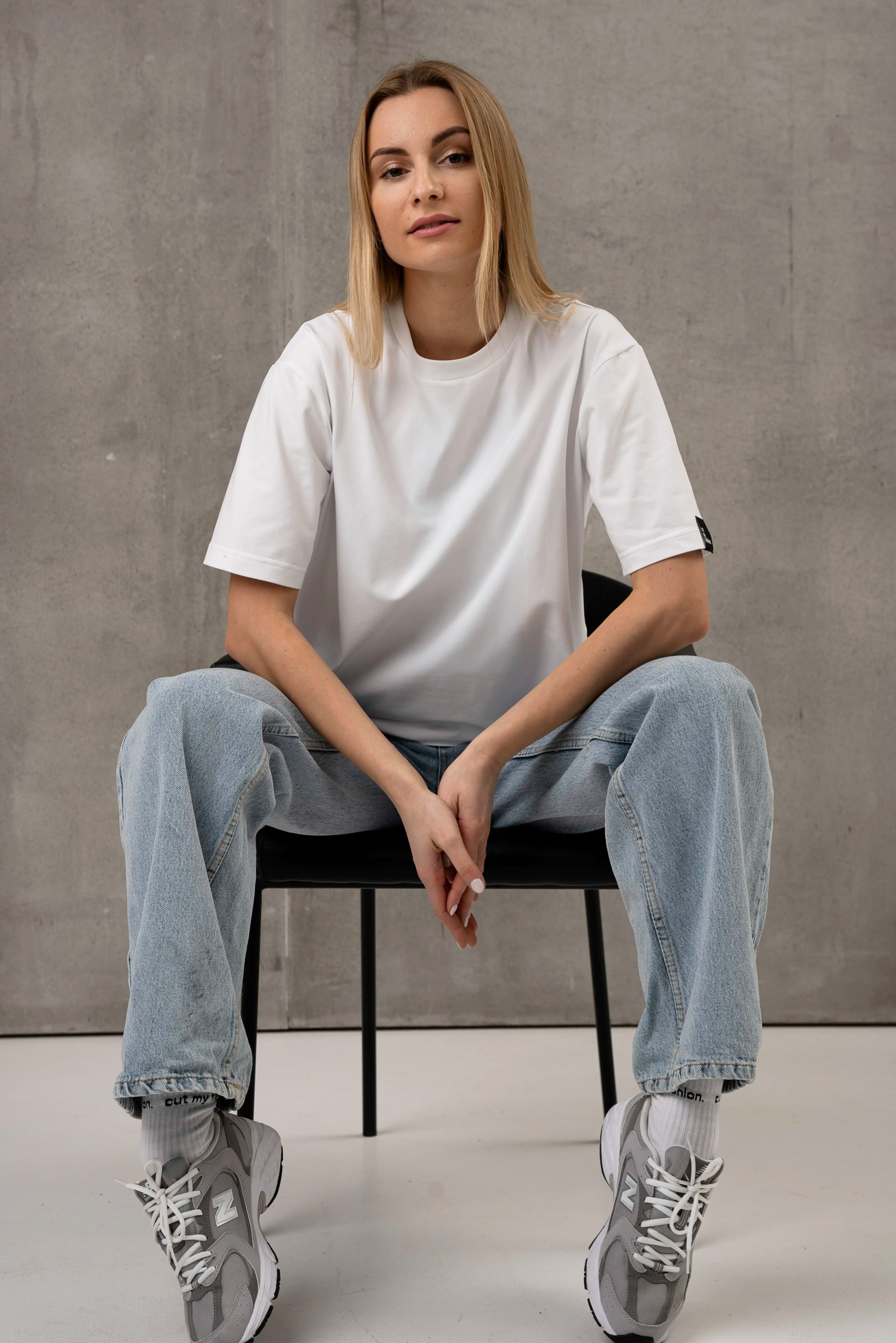 A woman is wearing a white, oversized short-sleeved T-shirt, light-colored jeans, and gray sneakers; she is sitting on a black chair against a gray background.