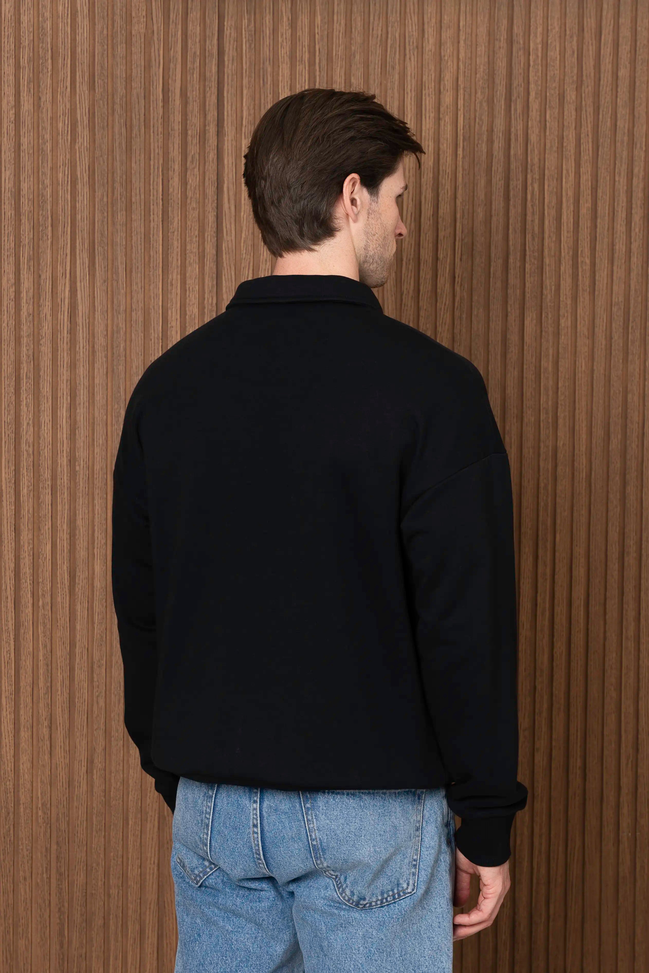 Vyras vilki nusisukęs nugarą juodą oversized džemperį su polo tipo apykakle ir trumpu sagų užsegimu priekyje, derintą su mėlynais džinsais.